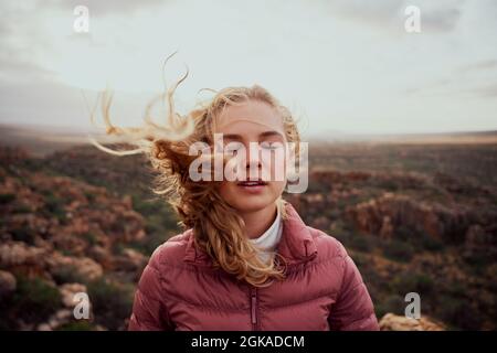 Giovane donna con occhi chiusi che sente vento fresco contro il viso in piedi sulla collina di montagna Foto Stock