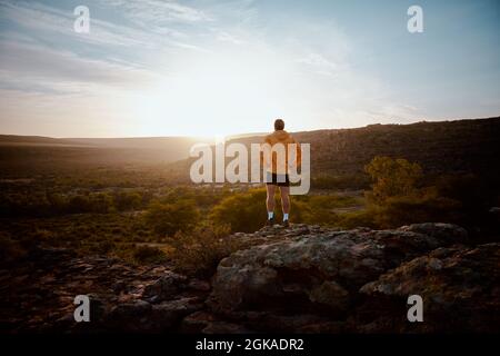 Vista posteriore del giovane atleta in piedi sulla scogliera di montagna dopo la corsa di mattina guardando la bella alba Foto Stock