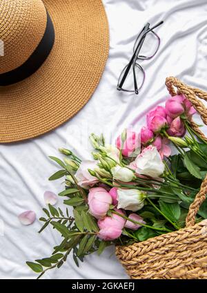 Cappello di paglia e bouquet di fiori rosa rosa su sfondo bianco. Vista dall'alto, composizione minima in stile di posa piatta. Concetto di vacanza estiva. Foto Stock