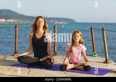 Mamma e figlia meditano su un molo vicino al mare, seduti su un tappeto in posizione lotus, in una serata estiva soleggiata Foto Stock
