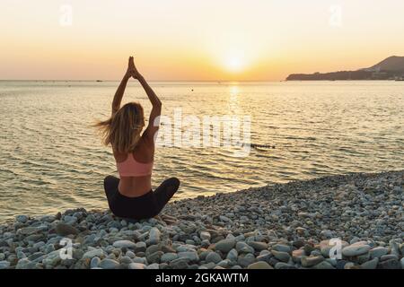 Una donna in abbigliamento sportivo che pratica yoga medita con le mani sollevate, seduto in posa di loto su una spiaggia di ciottoli in una serata estiva soleggiata. Foto Stock