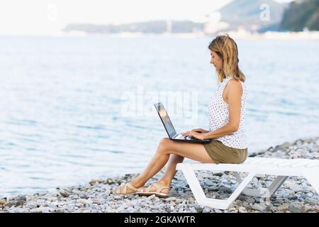 La donna freelance lavora da remoto mentre si siede su un lettino con un computer portatile sulla riva del mare Foto Stock