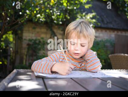 il bambino di cinque anni impara a leggere con la concentrazione. Si siede a un tavolo nel giardino. Abilità di lettura per i preschoolers Foto Stock