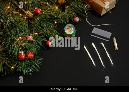 La cocaina suddivisa in percorsi con giocattoli di Natale e il ramo di abete rosso su sfondo nero in vista dall'alto. Foto Stock