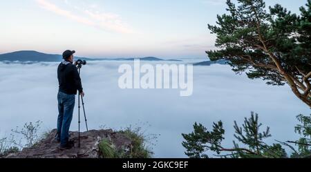 un fotografo uomo in montagna scatta foto con una macchina fotografica su un treppiede della natura e la nebbia mattutina delle colline. il primo tempo è l'alba e il mi Foto Stock