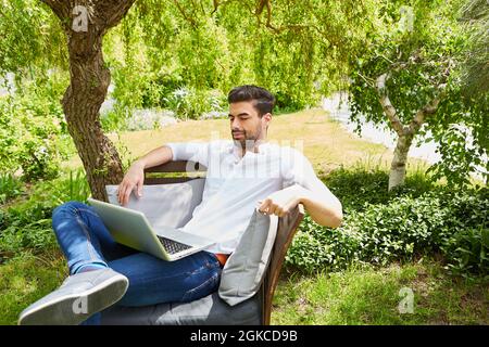 Il giovane come freelance lavora su un computer portatile nel giardino verde in estate Foto Stock