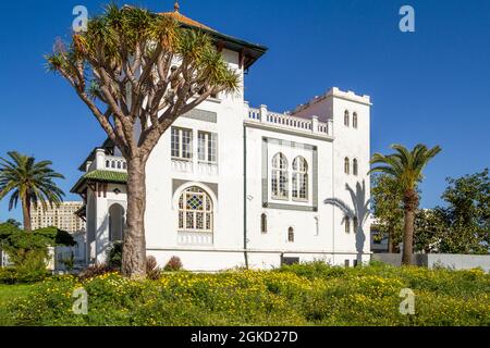 Edificio coloniale con una facciata bianca con piastrelle verdi circondate da alberi, palme e erba alta nella città di Tangeri, Marocco Foto Stock