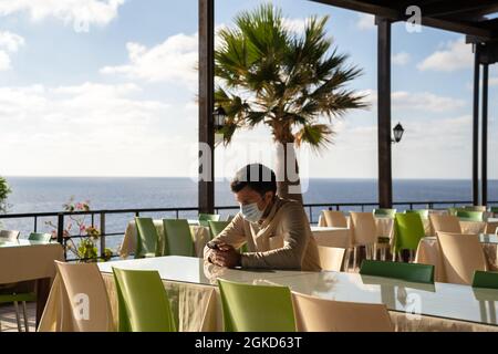 L'uomo mascherato solitario triste siede e usa lo smartphone in un ristorante vuoto a cipro ad Agios Georgios. Il visitatore indossa la maschera in attesa di ordine mentre è seduto Foto Stock