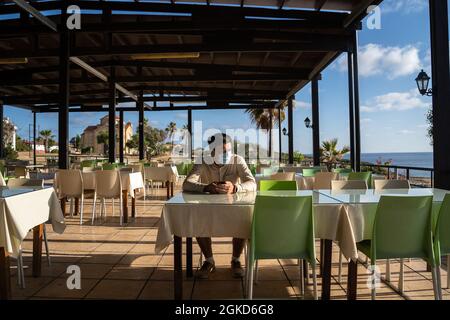 L'uomo mascherato solitario triste siede e usa lo smartphone in un ristorante vuoto a cipro ad Agios Georgios. Il visitatore indossa la maschera in attesa di ordine mentre è seduto Foto Stock