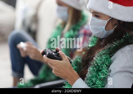 Donne in maschere mediche protettive e cappelli babbo natale seduti sul divano e tenendo console di gioco nelle loro mani a casa Foto Stock