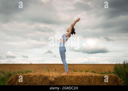 Donna caucasica che fa backbend in piedi su una balla di paglia in un campo, concetto di yoga di idoneità. Foto Stock