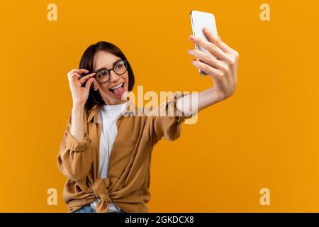 Donna giocosa che prende selfie, che mostra la lingua alla macchina fotografica del telefono cellulare e gli occhiali a contatto, che si posa su sfondo giallo Foto Stock