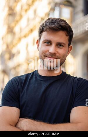 Sicuro positivo giovane bearded maschio in t shirt casual in piedi con le braccia incrociate contro l'edificio urbano sfocato e guardando la macchina fotografica nel giorno d'estate Foto Stock