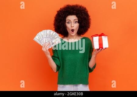 Donna scioccata con acconciatura afroche indossa il maglione verde casual stile in piedi con scatola regalo in mani e ventilatore di centinaia di dollari fatture. Monolocali al coperto Foto Stock