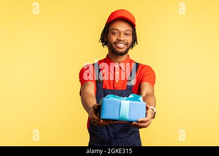 Un deliveryman sorridente con cappuccio rosso e uniforme blu che tiene fuori la scatola, consegna rapida, guardando la fotocamera sorridente. Monolocale interno Foto Stock