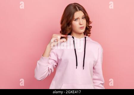 Ritratto di bruna dispiaciuta adolescente femmina in felpa con cappuccio facendo un po 'di gesto e guardando con volto insoddisfatto, mostrando minimo. Prigioniero interno Foto Stock