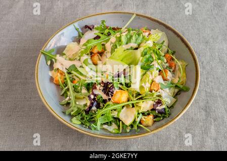 Ciotola di insalata verde con fette di mela, fette di formaggio, crostini, rucola e germogli di lattuga con salsa di maionese su tovaglia di lino grigio Foto Stock