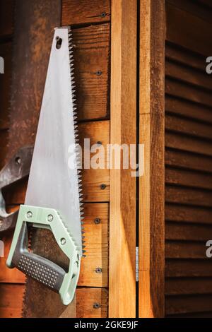 Una nuova sega a mano appende su un muro di legno in un laboratorio del villaggio vicino a vecchie seghe arrugginite, illuminate dalla luce del sole. Strumenti e vita rurale. Foto Stock