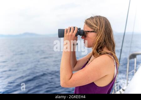 Giovane donna che guarda attraverso binocoli, in barca a vela, Dodecanesi, Grecia Foto Stock