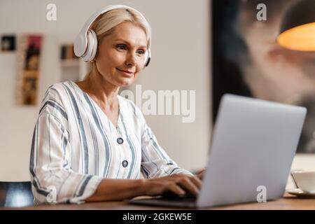 Donna bionda matura in cuffie sorridenti mentre lavora con il notebook a casa Foto Stock