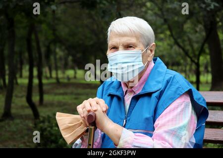 Donna anziana in maschera protettiva seduta con bastone da passeggio su una panca nel parco. Sicurezza durante la pandemia di coronavirus, vita in pensione Foto Stock