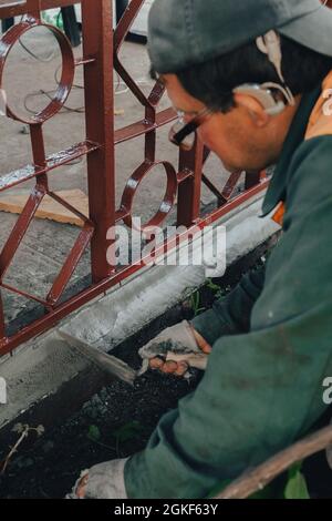 I tecnici edili con apparecchi acustici che lavorano con pavimentazione in cemento, si mescolano con la cazzuola sul lavoro laterale nel cortile. Polvere di Malta di cemento messa su mattone. REA Foto Stock