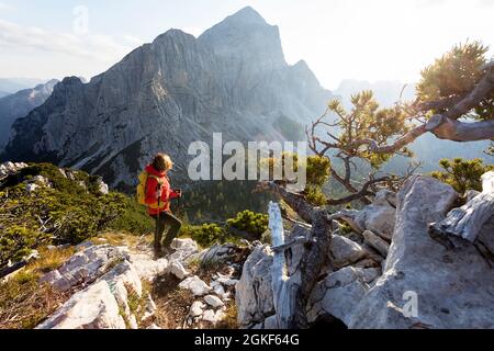 Donna in anorak rosso che cammina su un terreno roccioso sul crinale di montagna con una bella vista panoramica sulle alpi, Robicje, Alpi Giulie, Slovenia Foto Stock