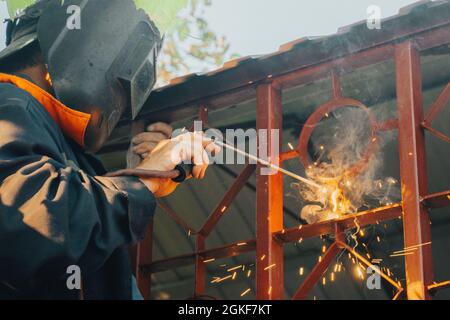 Primo piano di una saldatrice a mano saldante la parte metallica della struttura in acciaio nel cortile domestico. L'uomo dell'imbarcazione usa l'argon elettrico dell'arco di lavoro uniforme, casco protettivo o Foto Stock