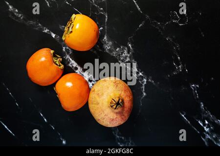 Immagine dall'alto di diversi persimmoni maturi con una deliziosa melograna su un tavolo in marmo nero Foto Stock