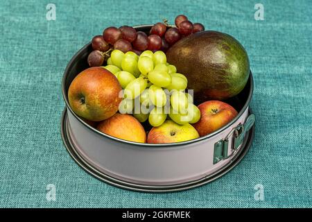 Ciotola di frutta rosa con mele, mango africano e grappoli di uva bianca e rossa su tessuto blu turchese Foto Stock