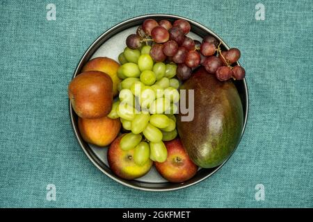 Immagine dall'alto di una ciotola di frutta con grappoli di uva rossa e bianca con mango maturo e mele su tela blu turchese Foto Stock