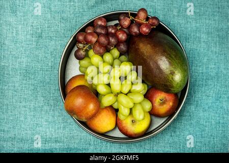 Immagine vista dall'alto di una ciotola di frutta con mele rosse, grappoli di uva bianca e rossa e mango africano su tovaglia blu Foto Stock