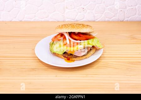 Hamburger speciale con molte fette di pomodoro, uova fritte che gocciolano il tuorlo sul prosciutto e vitello su un piatto bianco Foto Stock