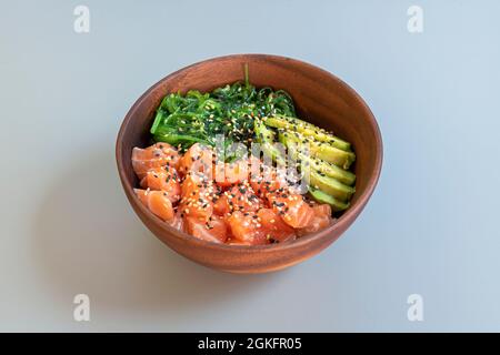 Ciotola di legno piena di sano poke di salmone con semi di papavero e sesamo, pezzi di avocado maturo e insalata di alghe di wakame su tavola blu Foto Stock