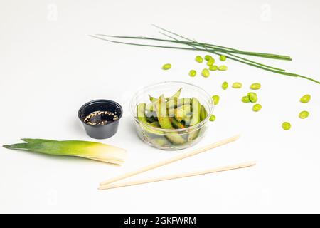 Cialde di edamame in recipiente di plastica trasparente con salsa di soia e fagioli di edamame su tavola bianca, chopstick monouso davanti. Foto Stock