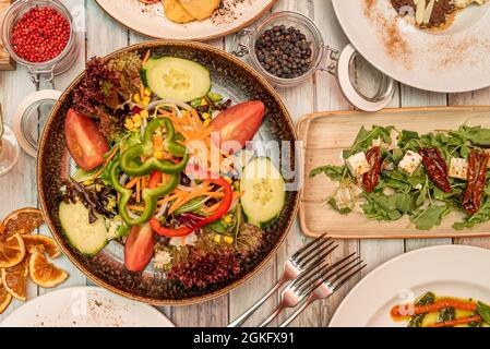 Immagine dall'alto di insalata di verdure e verdure fresche crude, insalata di rucola con formaggio feta e pomodori secchi, forchette, pepe rosso, pepe nero, arancio Foto Stock