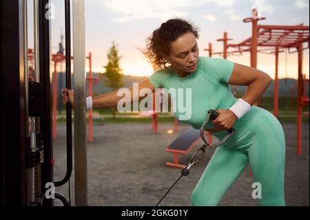 Bella atleta femminile che si esercita con la macchina da ginnastica Cable Crossover nella palestra all'aperto. Attraente fit donna che flette i muscoli sulla macchina da palestra. Foto Stock