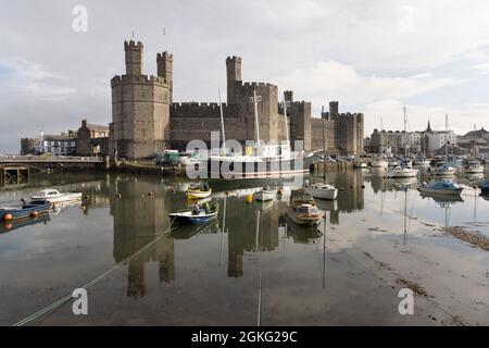 Caernarfon, Gwynedd, UK, 9 settembre 2021: Il castello di Edward i si riflette nel porto in cui sono ormeggiate piccole barche ed ex-nave luce Gran Bretagna. Foto Stock