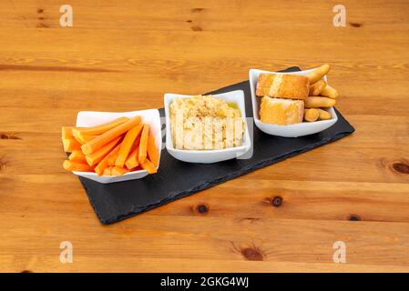 Hummus di ceci con olio d'oliva e sesamo accompagnati da strisce di carote, crostini e pane da immergere su un tavolo di legno Foto Stock