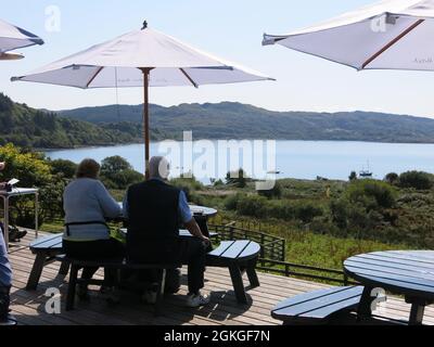 Vista posteriore di una coppia anziana seduta su una panca da picnic sotto un ombrellone che guarda sul Loch e ammira la vista in una giornata estiva soleggiata. Foto Stock