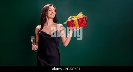 donna in abito nero contiene il regalo rosso scatola e bicchiere di champagne. Ora di festa. Concetto di vacanza Foto Stock