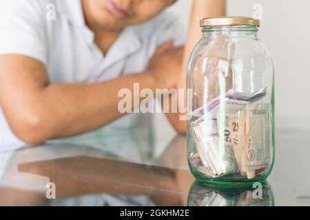 la mano del ragazzo accanto ai suoi risparmi in un vaso di vetro. il giovane uomo accanto alle sue finanze personali. annoiato studente con una mano sulla testa. risparmi e finanza con Foto Stock