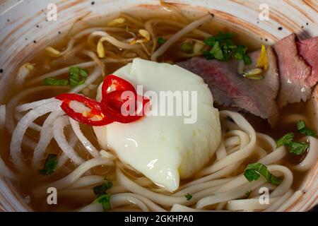 Zuppa di pho bo in stile asiatico con spaghetti di udon, uova in camicia, funghi e cipolla verde in un recipiente sul tavolo. Vista dall'alto orizzontale Foto Stock