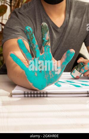 dettagli dello spazio di lavoro di un artista che mostrano le palme delle sue mani macchiate con vernice blu chiaro, su una scrivania con un libro di esercizi, stile di vita Foto Stock