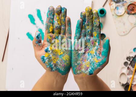 dettagli dello spazio di lavoro di un artista che mostra le palme delle sue mani macchiate di vernice, su una scrivania con vari strumenti di lavoro come tavolozze, pennelli, carta Foto Stock