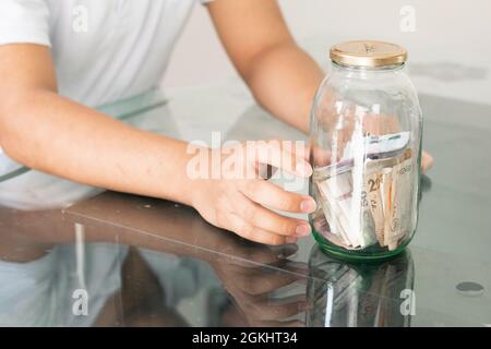 mano del bambino che raccoglie i loro risparmi in un vaso di vetro. giovane uomo vicino alle sue finanze personali. ragazzo frettoso. risparmio e concetto di finanza Foto Stock