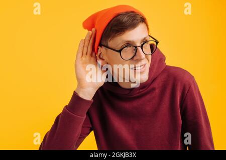 l'uomo bello è duro di udito, in piedi su uno sfondo giallo, tiene la mano vicino al suo orecchio, overhears, concetto di non udente Foto Stock