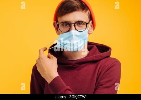 bell'uomo in una maschera protettiva con occhiali e un cappello arancione e una felpa rossa, toglie la sua maschera protettiva, su sfondo giallo, felice va Foto Stock