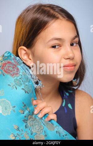 Primo piano ritratto di una ragazza adolescente dai capelli scuri e carini di 12 anni in un abito blu scuro, lei scherzava giocosamente fuori da dietro un cuscino di seta blu. Foto Stock