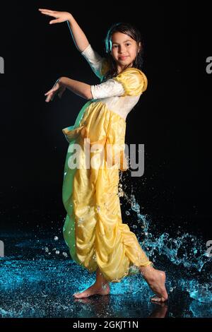 Ragazza adolescente dai capelli scuri, bella e allegra, snella, di 12 anni in un abito di seta giallo a piedi nudi in un aquazone nero. Emozioni e danza dentro Foto Stock